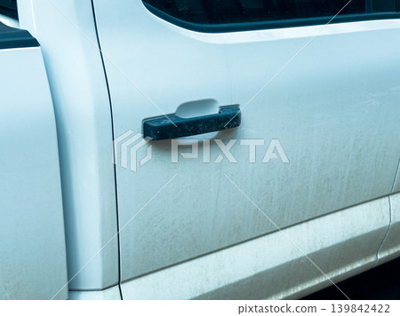 Dust covers the door handle of a white pickup truck parked in a lot during daylight hours in a suburban area with no visible people Dust covers the door handle of a white pickup truck parked in a lot during daylight hours in a suburban area with no visible people 139842422