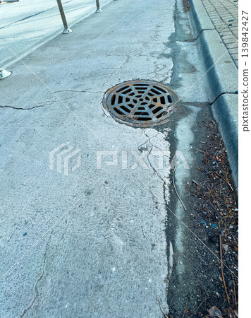 Street view showing cracked pavement and a storm drain cover on a city sidewalk during daytime 139842427