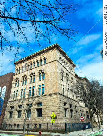 Historic stone building stands tall in urban setting under blue sky with light clouds and bare tree branches on a clear day 139842428
