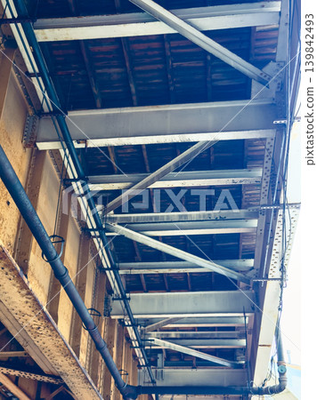 View of a train bridge structure showing metal beams and wooden supports under a rail line in a city during daylight hours View of a train bridge structure showing metal beams and wooden supports under a rail line in a city during daylight hours 139842493