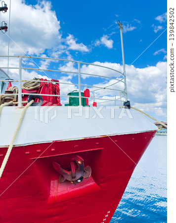 Red ship docked at the harbor with ropes, anchor, and green objects on deck under a blue sky with clouds Red ship docked at the harbor with ropes, anchor, and green objects on deck under a blue sky with clouds 139842502