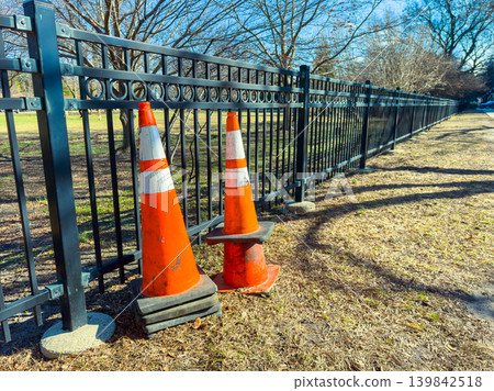 Two orange traffic cones stand beside a black metal fence in a park during a clear daytime scene Two orange traffic cones stand beside a black metal fence in a park during a clear daytime scene 139842518