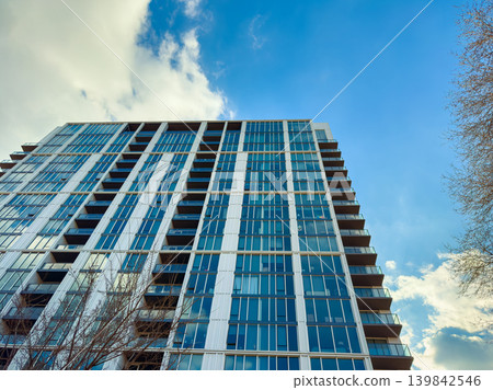 View of a tall modern building with large glass windows under a blue sky in a city during the daytime View of a tall modern building with large glass windows under a blue sky in a city during the daytime 139842546