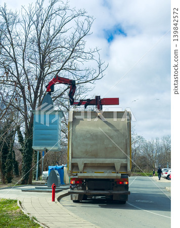 Waste collection truck uses a mechanical arm to lift a blue container in a busy street during the day while trees are bare in early spring 139842572