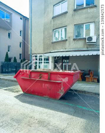 Construction waste bin placed on a street near a building during the day with shadows and nearby furniture visible Construction waste bin placed on a street near a building during the day with shadows and nearby furniture visible 139842573