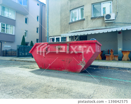Construction work takes place near a red dumpster placed outside a building on a city street during daytime 139842575