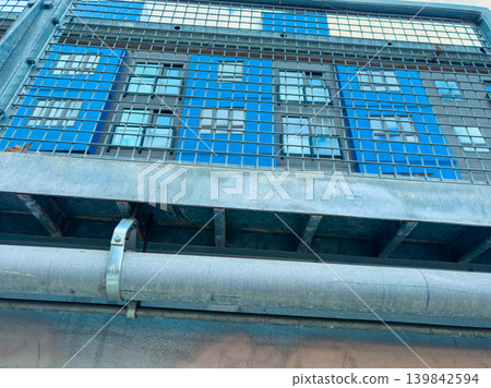 View of a building with blue walls seen through a metal grid during daytime in an urban area with clear skies and sunlight View of a building with blue walls seen through a metal grid during daytime in an urban area with clear skies and sunlight 139842594