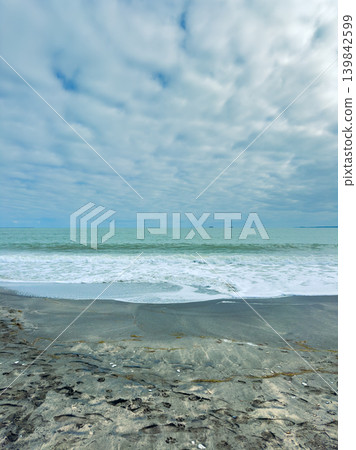 View of the ocean waves rolling onto the shore under a cloudy sky at a coastal location in the afternoon View of the ocean waves rolling onto the shore under a cloudy sky at a coastal location in the afternoon 139842599