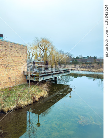Wooden dock on a pond near a brick building with trees and cloudy sky in a quiet area during late afternoon Wooden dock on a pond near a brick building with trees and cloudy sky in a quiet area during late afternoon 139842624