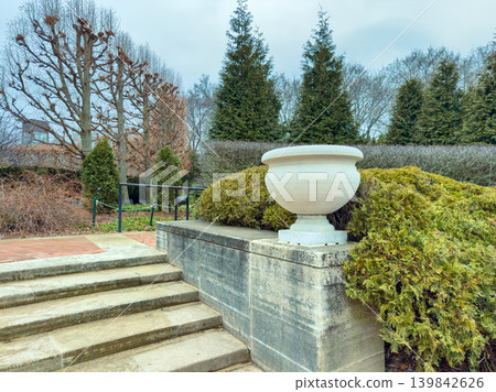 Steps lead to a garden with a large stone pot and trimmed bushes near bare trees on a cloudy day 139842626