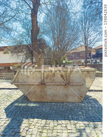 Large metal container placed on cobblestone road under bare trees across from buildings on a sunny day Large metal container placed on cobblestone road under bare trees across from buildings on a sunny day 139842639