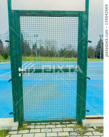 Green gate leads to empty tennis court in a park during a cloudy day with grass and trees in the background Green gate leads to empty tennis court in a park during a cloudy day with grass and trees in the background 139842669