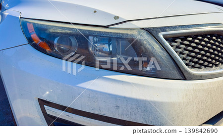 White car parked under sunlight shows details of front headlight and grime indicating lack of recent cleaning White car parked under sunlight shows details of front headlight and grime indicating lack of recent cleaning 139842690
