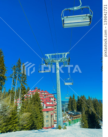 Snow-covered mountain ski resort with chairlift in bright blue sky during sunny day in winter showcasing ski activities and nearby buildings Snow-covered mountain ski resort with chairlift in bright blue sky during sunny day in winter showcasing ski activities and nearby buildings 139842721