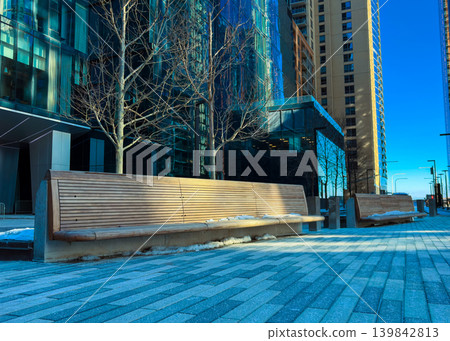 People walk on the path near benches surrounded by tall buildings and trees in a city on a sunny day in winter People walk on the path near benches surrounded by tall buildings and trees in a city on a sunny day in winter 139842813