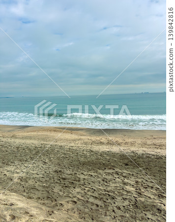 Ocean waves crash on sandy beach at midday under cloudy sky near coastline with distant ships on horizon Ocean waves crash on sandy beach at midday under cloudy sky near coastline with distant ships on horizon 139842816