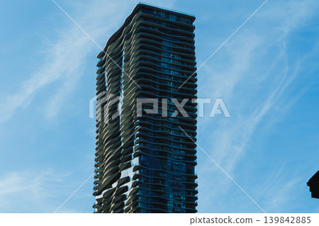 Tall building stands against blue sky in a city during daytime with clouds and sunlight highlighting its structure Tall building stands against blue sky in a city during daytime with clouds and sunlight highlighting its structure 139842885