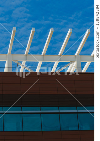 Focus on modern architecture with white beams and large glass windows under a blue sky in a city setting during daylight hours Focus on modern architecture with white beams and large glass windows under a blue sky in a city setting during daylight hours 139842894