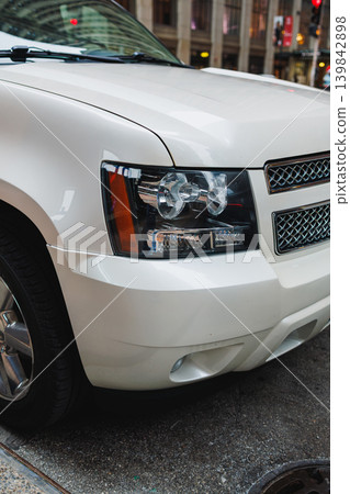 White SUV parked on a city street near tall buildings, showcasing the front view with headlights and chrome grille on a busy afternoon in the urban setting White SUV parked on a city street near tall buildings, showcasing the front view with headlights and chrome grille on a busy afternoon in the urban setting 139842898