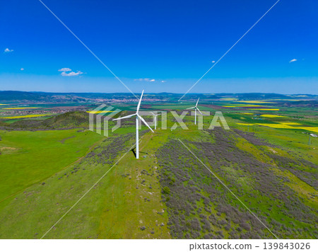 Wind turbines stand tall in a green field under a clear blue sky near a small town at midday in early spring Wind turbines stand tall in a green field under a clear blue sky near a small town at midday in early spring 139843026