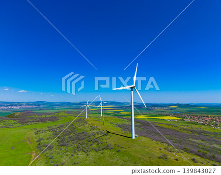 Wind turbines stand on a green hill under a blue sky in the countryside with fields and small buildings in the distance on a sunny day Wind turbines stand on a green hill under a blue sky in the countryside with fields and small buildings in the distance on a sunny day 139843027
