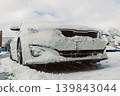 Snow covers a parked car on a winter day in a residential area with houses and trees visible in the background 139843044
