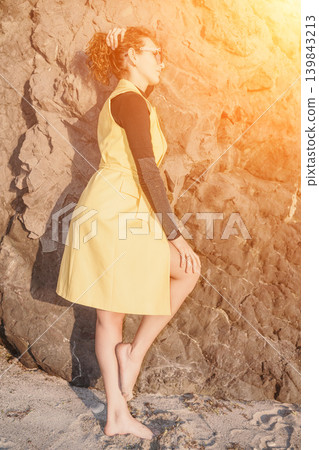 Woman yellow dress beach posing against a rocky cliff wall during a warm golden hour sunset with lens flare Woman yellow dress beach posing against a rocky cliff wall during a warm golden hour sunset with lens flare 139843213