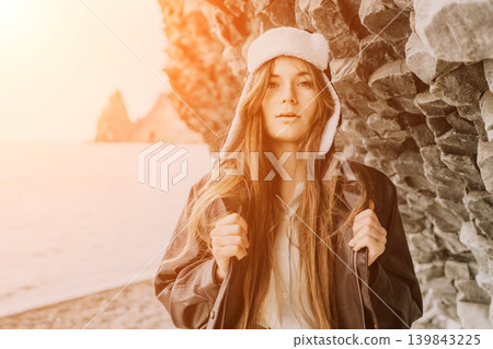 Woman beach winter wearing a warm hat and jacket stands on a pebbly shore during a golden sunset with copy space 139843225
