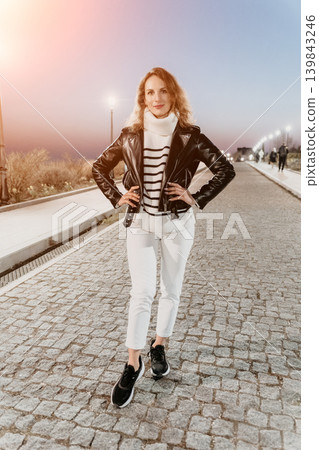 Woman Streetwear Evening stylish woman wearing a leather jacket and white jeans poses on a cobblestone walkway at sunset Woman Streetwear Evening stylish woman wearing a leather jacket and white jeans poses on a cobblestone walkway at sunset 139843246