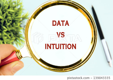 A hand holds a magnifying glass over a white surface, revealing the words Data versus Intuition. A sleek pen rests nearby, beside a small green plant 139843335