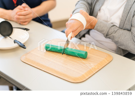 A senior man undergoing rehabilitation and cooking training using a kitchen knife, along with an occupational therapist and care manager. 139843859