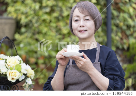 一位老婦人在自家院子裡一邊喝咖啡一邊種花。 139844195