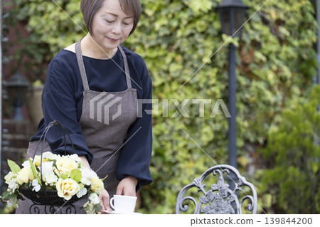 一位老婦人在自家院子裡一邊喝咖啡一邊種花。 一位老婦人在自家院子裡一邊喝咖啡一邊種花。 139844200