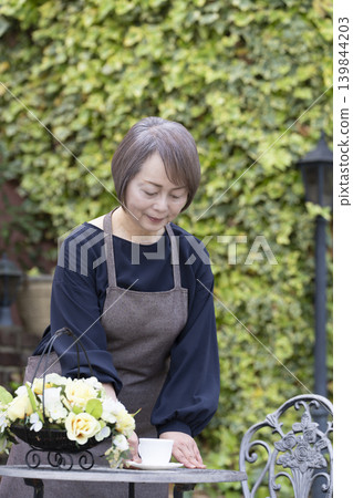 一位老婦人在自家院子裡一邊喝咖啡一邊種花。 一位老婦人在自家院子裡一邊喝咖啡一邊種花。 139844203