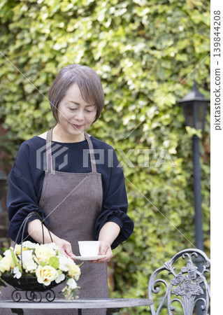 一位老婦人在自家院子裡一邊喝咖啡一邊種花。 一位老婦人在自家院子裡一邊喝咖啡一邊種花。 139844208