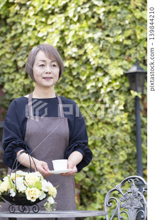 一位老婦人在自家院子裡一邊喝咖啡一邊種花。 一位老婦人在自家院子裡一邊喝咖啡一邊種花。 139844210