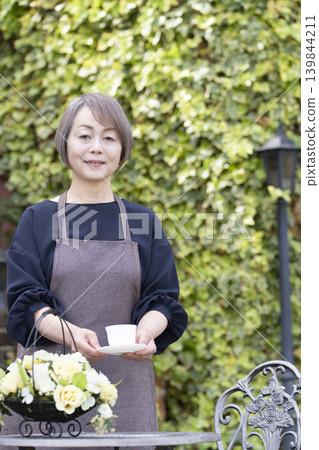 A senior woman drinking coffee while gardening in her yard. A senior woman drinking coffee while gardening in her yard. 139844211