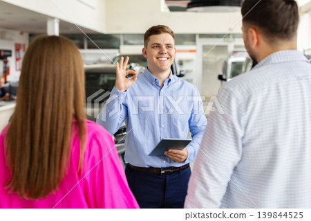 Friendly car salesman showing approval gesture while consulting couple in dealership showroom Friendly car salesman showing approval gesture while consulting couple in dealership showroom 139844525
