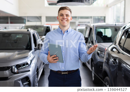 Smiling car salesman with tablet standing between vehicles at auto dealership showroom Smiling car salesman with tablet standing between vehicles at auto dealership showroom 139844527