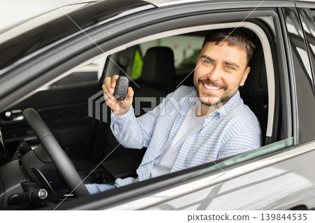 Happy young man sitting in new car at dealership holding key fob Happy young man sitting in new car at dealership holding key fob 139844535