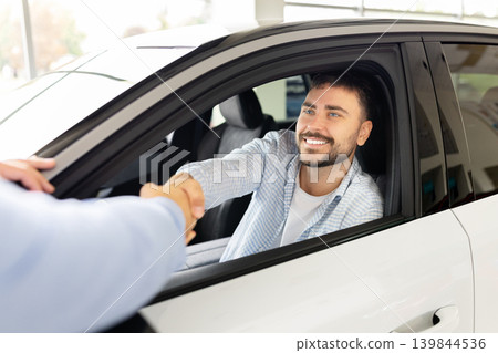 Young male customer shaking hands with professional salesman from inside new automobile Young male customer shaking hands with professional salesman from inside new automobile 139844536