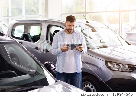 Man using digital tablet while comparing vehicles inside modern car dealership showroom Man using digital tablet while comparing vehicles inside modern car dealership showroom 139844549