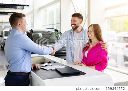 Happy couple shaking hands with car salesman at dealership after vehicle purchase Happy couple shaking hands with car salesman at dealership after vehicle purchase 139844552