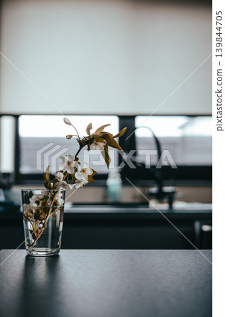 Moody minimalist kitchen interior with white cherry blossoms in glass. Modern dark counter with blurred sink background. Elegant spring branch symbolizing home wellness and slow living aesthetic. Moody minimalist kitchen interior with white cherry blossoms in glass. Modern dark counter with blurred sink background. Elegant spring branch symbolizing home wellness and slow living aesthetic. 139844705