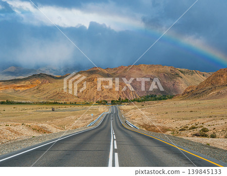 An empty asphalt road stretches through dry Altai mountains. A colorful rainbow brightens the dark, cloudy sky after rain An empty asphalt road stretches through dry Altai mountains. A colorful rainbow brightens the dark, cloudy sky after rain 139845133