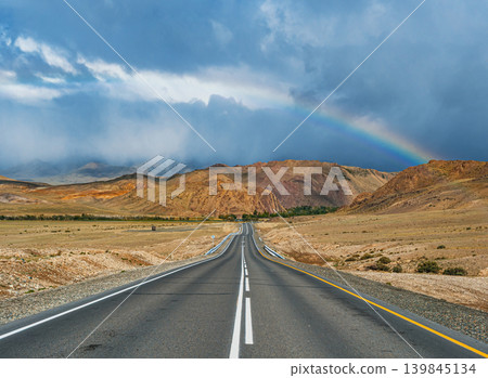 An empty asphalt road stretches through dry Altai mountains. A colorful rainbow brightens the dark, cloudy sky after rain An empty asphalt road stretches through dry Altai mountains. A colorful rainbow brightens the dark, cloudy sky after rain 139845134
