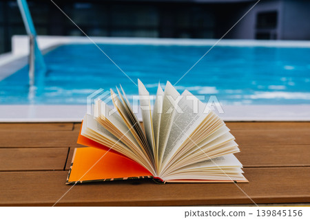 Open book on wooden poolside deck with blue swimming pool background. Summer reading, vacation learning and relaxation. Leisure travel lifestyle Open book on wooden poolside deck with blue swimming pool background. Summer reading, vacation learning and relaxation. Leisure travel lifestyle 139845156