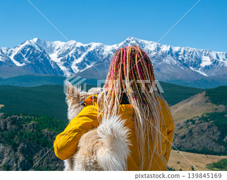 A person with colorful dreadlocks and a yellow jacket holds a fluffy dog, looking out at the vast Altai mountains A person with colorful dreadlocks and a yellow jacket holds a fluffy dog, looking out at the vast Altai mountains 139845169