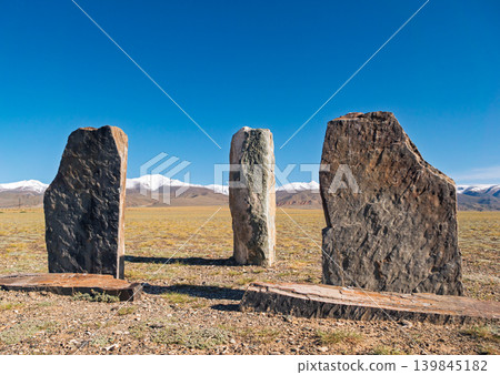 Three ancient stone monuments stand tall on a dry Altai plain. Distant snow capped mountains rise under a clear blue sky 139845182
