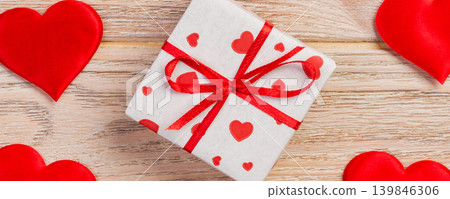 Valentine or other holiday handmade present in paper with red hearts and gifts box in holiday wrapper. Present box of gift on orange wooden table top view with copy space, empty space for design Valentine or other holiday handmade present in paper with red hearts and gifts box in holiday wrapper. Present box of gift on orange wooden table top view with copy space, empty space for design 139846306